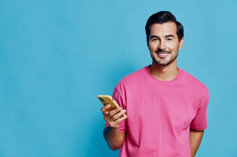 Smiling man wearing pink t-shirt, holding mobile phone with cheerful expression against bright blue background, casual lifestyle concept.