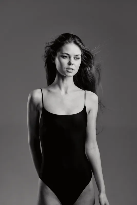 Black and white portrait of a slender woman in a black swimsuit with a calm expression on a plain background