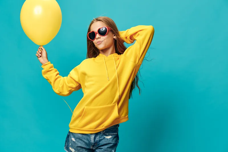 Girl wearing yellow hoodie and sunglasses holding yellow balloon posing against blue background for lifestyle, fashion and casual style concept with confident expression.