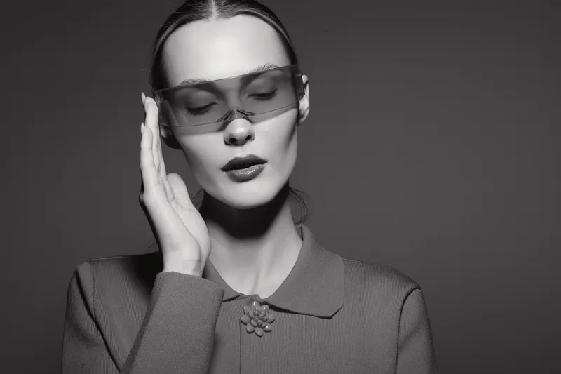 Young white woman with pale skin wearing futuristic translucent glasses and vintage style coat poses with closed eyes in monochrome studio portrait.