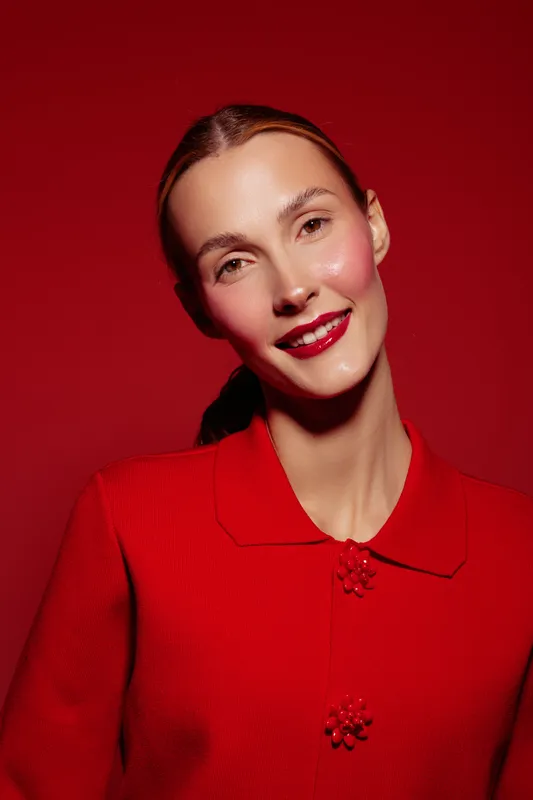 Smiling woman with glossy red lips and natural makeup poses against a red background wearing a red sweater. Concept of beauty, fashion, and confident female portrait in studio lighting.