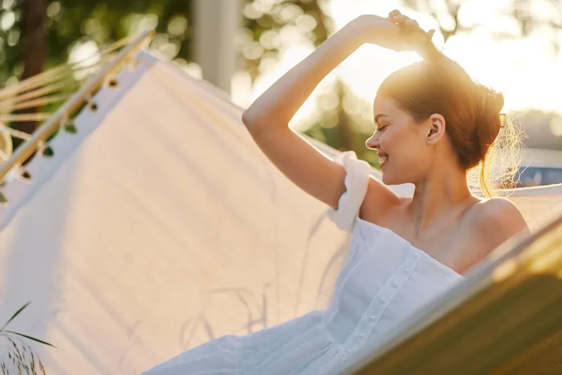 Young woman in a white dress relaxing in a hammock during sunset, radiating joy and tranquility amidst a serene outdoor setting