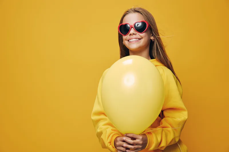 Girl smiling and holding yellow balloon on bright yellow background wearing sunglasses and casual sweater for happy carefree mood and fun portrait.