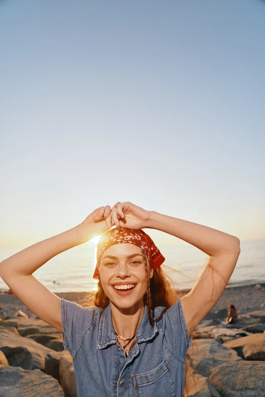 Lifestyle woman in modern boho-western denim street style with joyful expression, wearing a red bandana, enjoying the golden hour on the beach with warm film color tones.