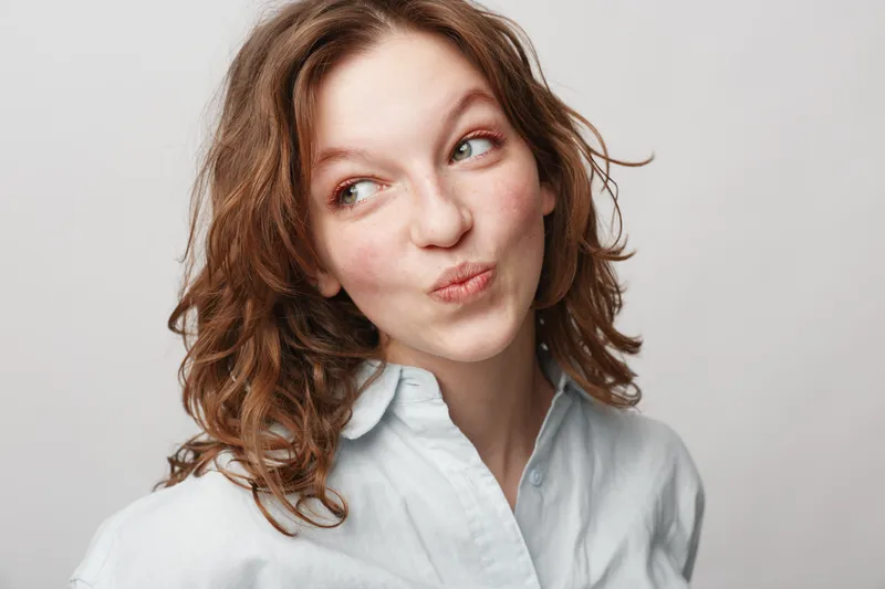 Young woman with curly hair wearing white shirt looks sideways with playful expression on light background. Concept of casual style, emotions, and natural beauty portrait.