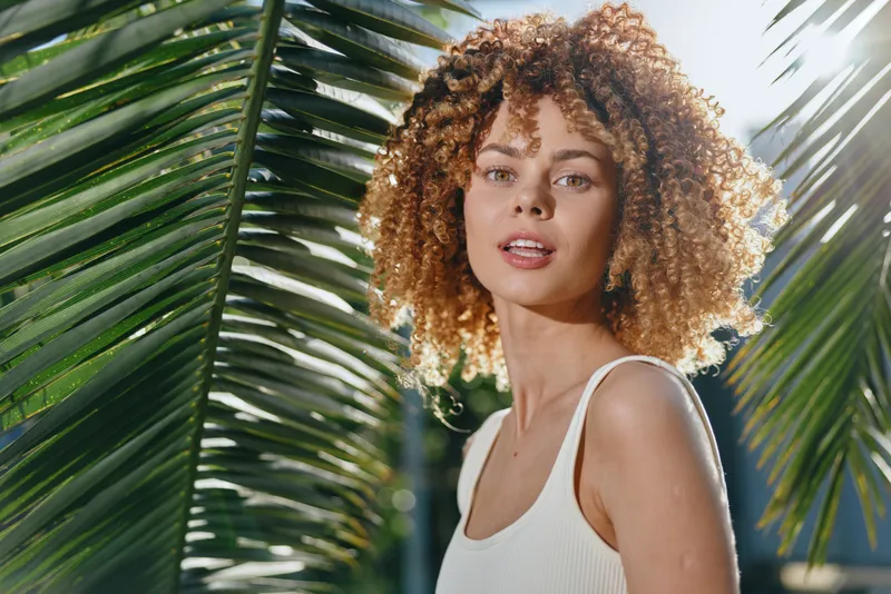 young woman with curly hair stands outdoors near green palm leaves under bright sunlight, wearing white tank top, radiating natural beauty and summer vibes