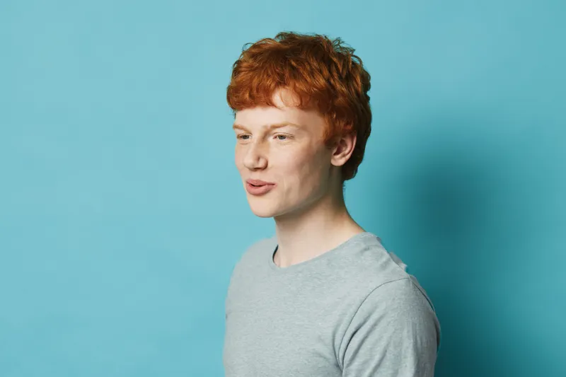 Young redhead man wearing gray shirt looking sideways with thoughtful expression against blue background for portrait and casual style concept.