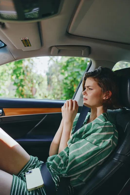 Young woman sits in a car interior, wearing a green striped shirt, looking thoughtful as she rests her hand near her mouth.