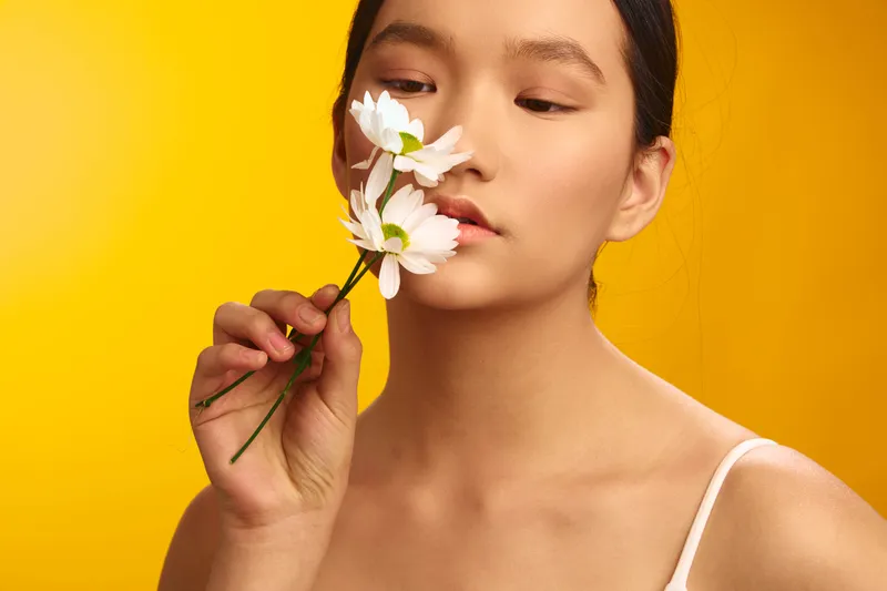 asian woman holds white flowers near her face against a bright yellow backdrop, presenting a serene lifestyle portrait that blends natural beauty with gentle mood and contemporary style.