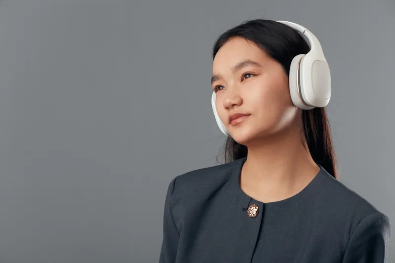 Woman with headphones listens to music or audio content, looking thoughtful and calm with neutral gray background and casual dark clothing in studio portrait.