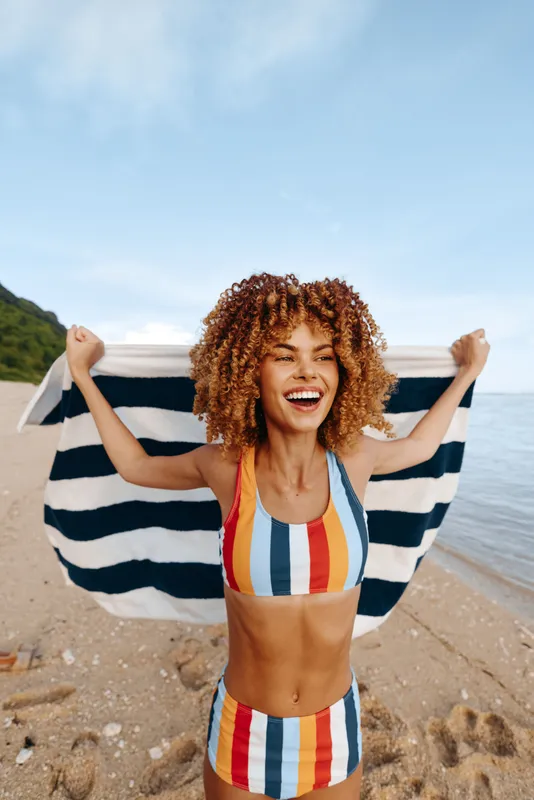 Smiling woman on a sandy beach holds a striped towel behind her shoulders, wearing a colorful striped swimsuit. Sunlit seaside scene exudes summer joy and relaxed wellness.