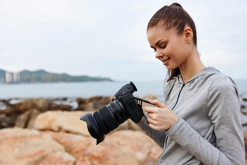 Young female photographer checking her camera by the seaside, wearing a grey hoodie, with a serene expression and a coastal background