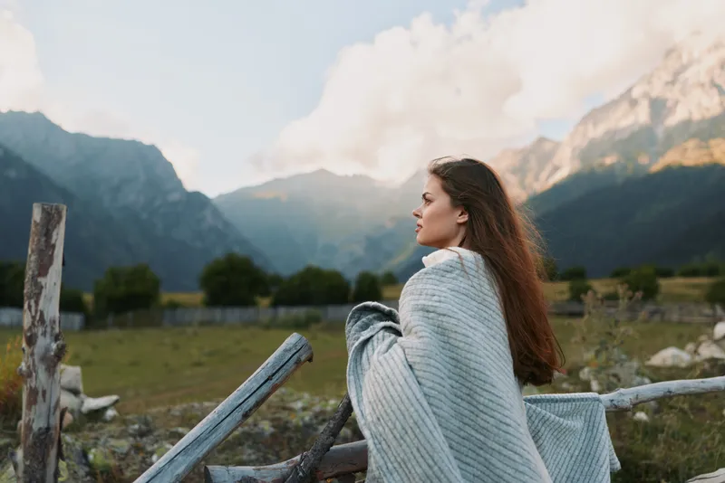 Young woman wrapped in blanket stands near wooden fence looks at mountain landscape enjoys peaceful outdoor lifestyle in nature during cloudy day.