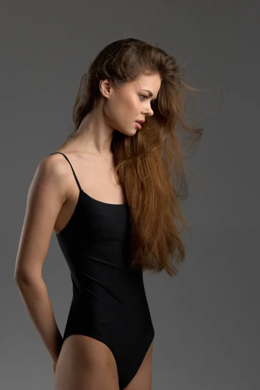 Profile of slender woman in black swimsuit with loose hair posing over gray background