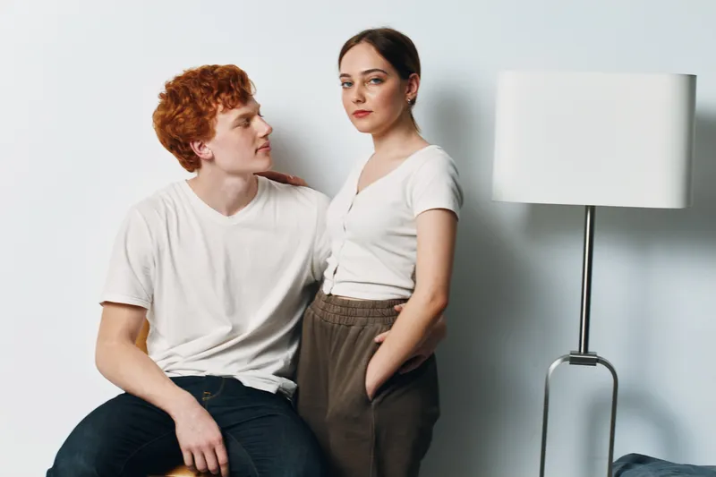 Young couple, man and woman, posing indoors against white wall in casual clothes. They stand close with serious expressions showing connection and relationship in minimalistic room with lamp.
