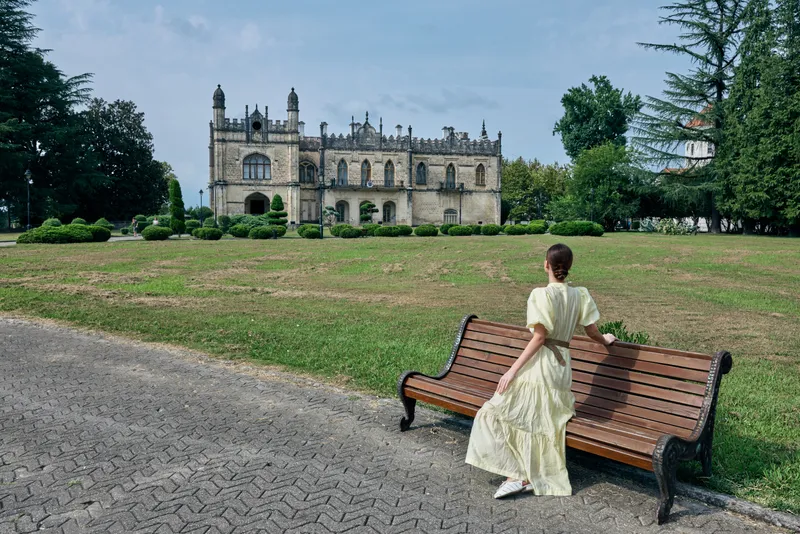 A woman in a vintage dress sits on a wooden bench in a park, facing a grand historic castle, capturing a quiet moment of timeless elegance and scenic architecture.