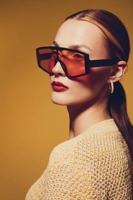 Stylish woman with long hair wearing oversized orange sunglasses and hoop earrings poses against a warm yellow background. Fashion portrait with modern accessories and confident expression.