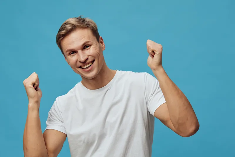 cheerful smiling young man wearing casual white t-shirt celebrating success with fists up isolated blue background studio portrait. People happiness concept