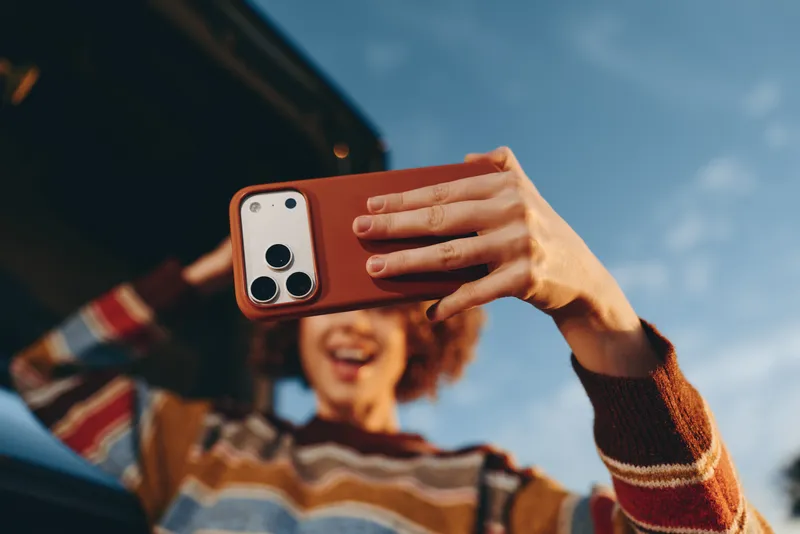 Woman lifestyle selfie outdoors, sunlit hands holding smartphone, smiling young woman blurred in background, car nearby, casual knit sweater, relaxed lifestyle moment, blue sky, daylight