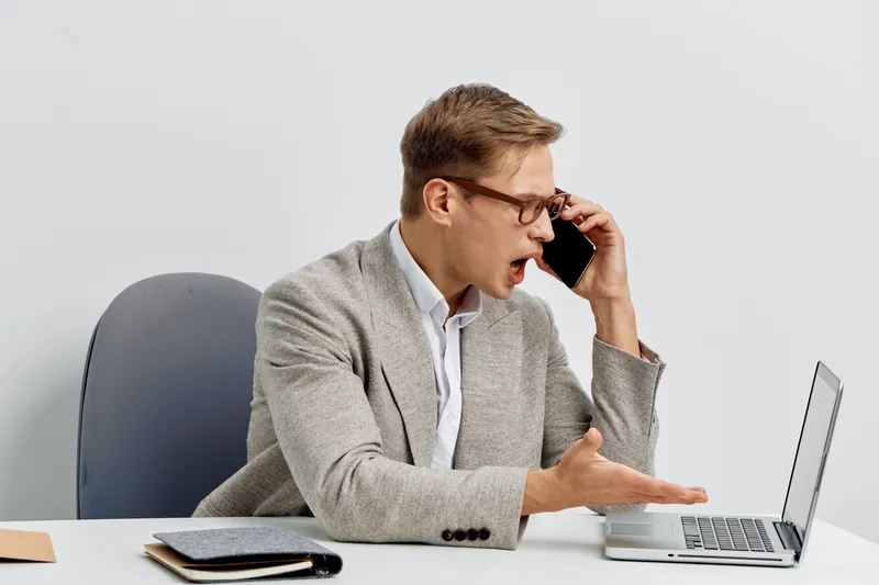 Frustrated businessman talking on mobile phone during work at office desk, reacting emotionally to unexpected news, wearing glasses and beige blazer, modern workplace environment, concept of workplace