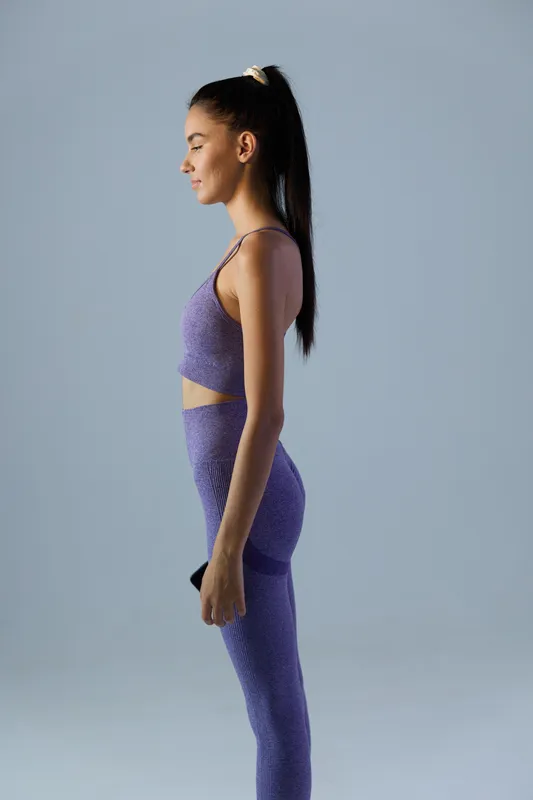 woman sport fitness in purple activewear stands calm and focused in profile, young athletic adult with ponytail against plain gray background, healthy lifestyle concept
