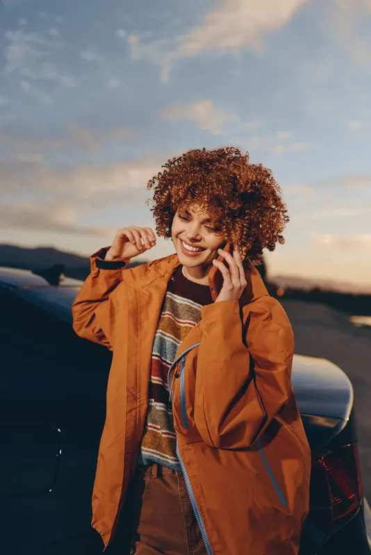 Woman lifestyle travel outdoors sunset curly hair jacket smiling sky, smiling woman adjusts her curly hair and poses near an airplane at golden hour, cinematic clouds and warm light.