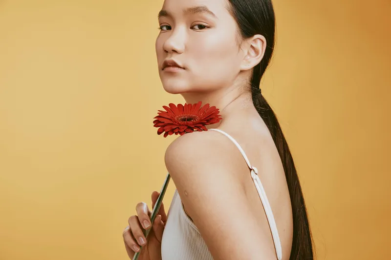 Asian woman gracefully poses with a red flower near her shoulder, set against a warm amber backdrop, evoking elegance, poise, softness, and contemporary portrait photography artistry.