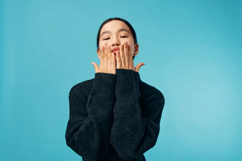 Woman with black sweater blowing a kiss with both hands to camera on blue background. Female portrait with closed eyes and playful facial expression in studio lighting.