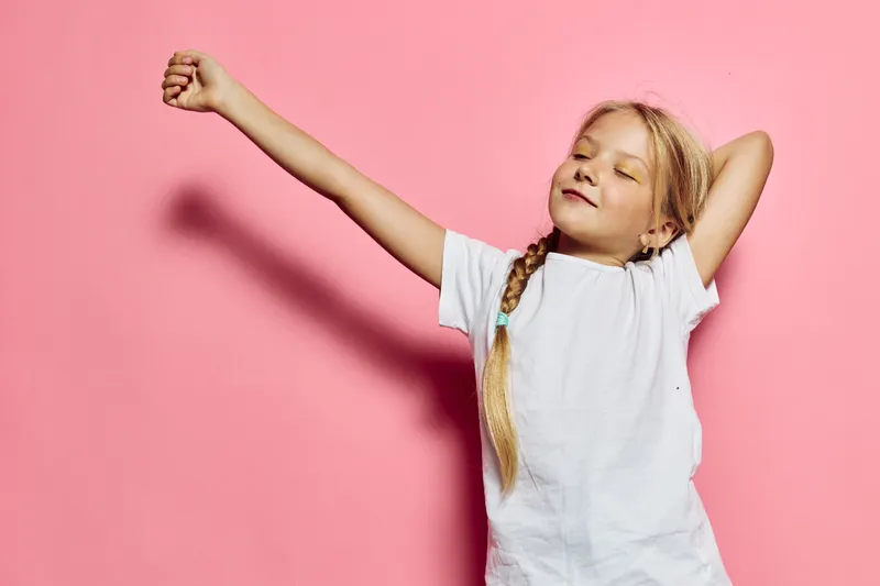 Young girl with long braided hair stretching her arms against a pink background. She wears a white t-shirt and expresses relaxation and comfort. Studio portrait.