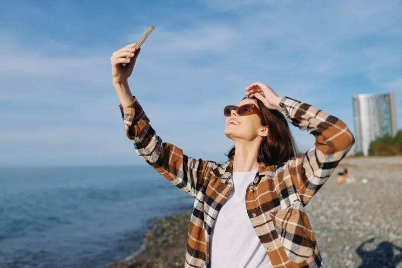 Woman by the seaside taking a selfie with a smartphone, outdoors on a sunny day, wearing a plaid shirt and sunglasses, ocean beside her with distant urban silhouettes.