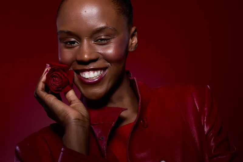 Smiling African American woman with dark skin holds red rose near face. She wears red leather jacket and stands against red background. Concept of beauty, happiness, and floral elegance.
