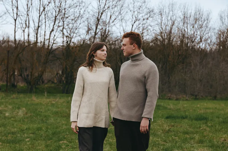 Couple outdoors wearing knit sweaters, calm conversation, soft light, natural colors, grassy field with bare trees, cozy fashion moment, candid connection, relaxed mood, seasonal ambiance.