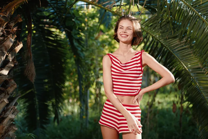 A cheerful young woman in a striped red and white swimsuit stands in a sunny tropical garden, smiling brightly as palm leaves frame the scene.