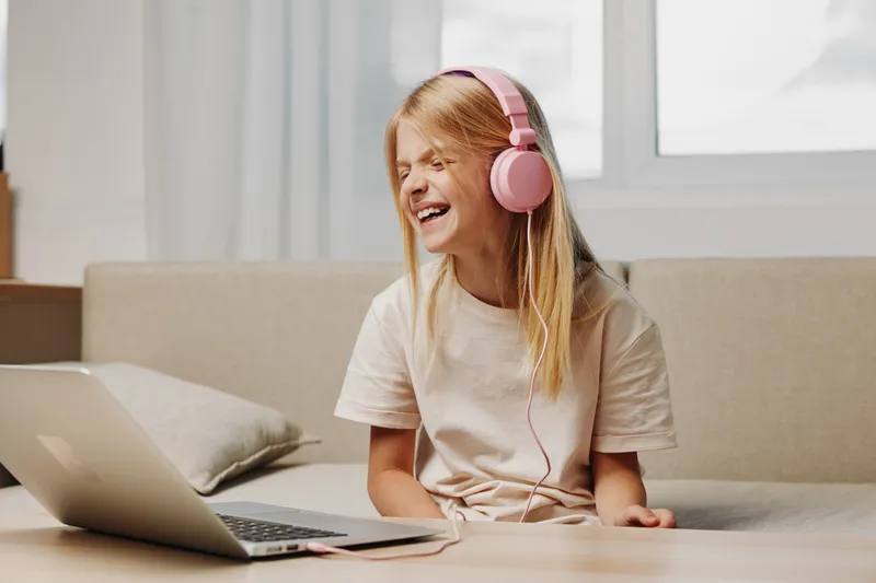 Happy girl with long hair wearing pink headphones enjoys music while sitting on couch with laptop. Bright room with natural light creates cheerful relaxed mood.
