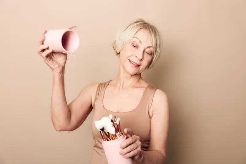 Happy mature woman with short hair holding pink containers and makeup brushes, smiling gently on beige background. Concept for beauty routine, skincare, and self-care at any age.