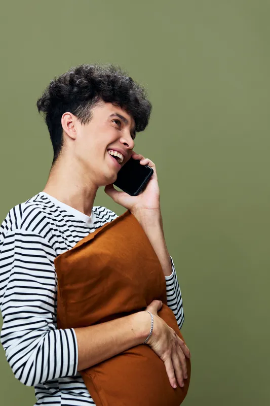 smiling man talking on smartphone holding orange pillow against green background, casual clothing, relaxed mood, indoor portrait, modern lifestyle