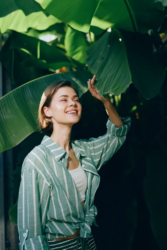 A young woman stands among large tropical leaves, smiling softly as sunlight filters through the greenery.