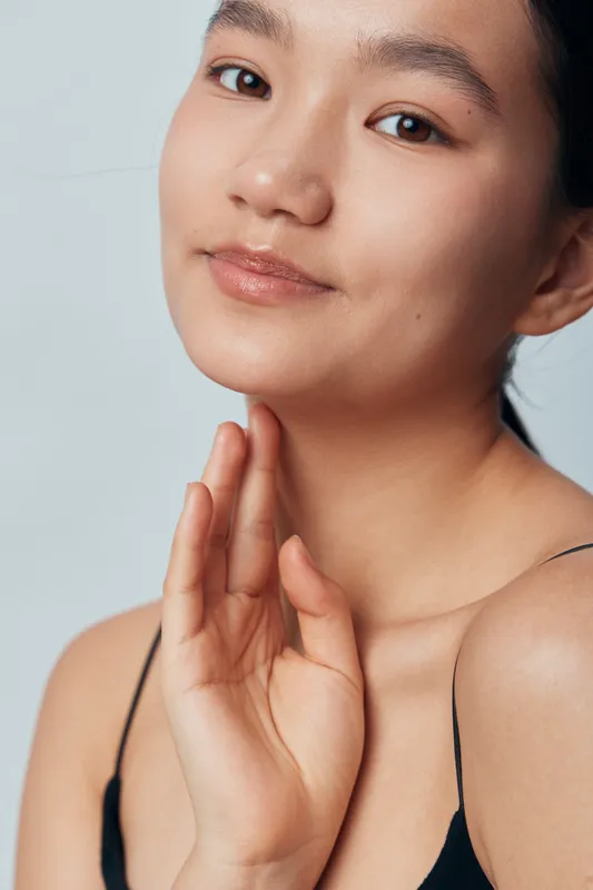 Woman with smooth skin and natural makeup posing in a black strap top, showing hand near face, beauty portrait with soft expression and gentle lighting on light background.