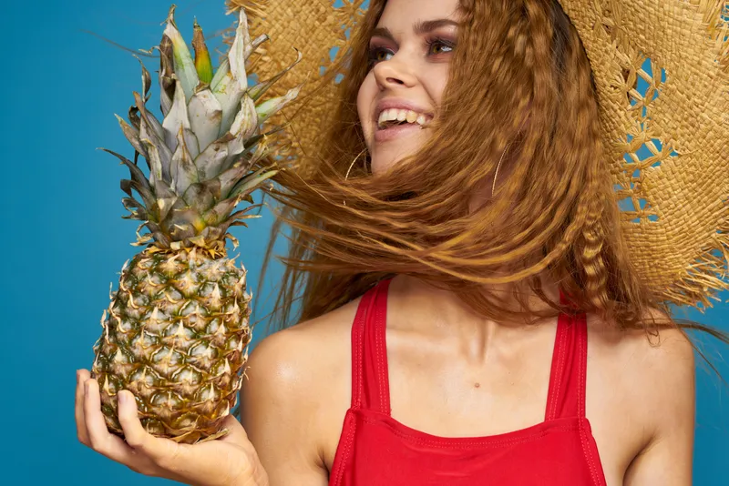 Женщина в соломенной шляпе держит в руках ананас на фоне синих экзотических фруктов