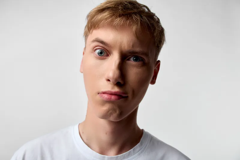 Close up of a puzzled young man wearing a white tee against a soft light background, raised eyebrow and a tilted head conveying confusion and curiosity in a clean studio setup