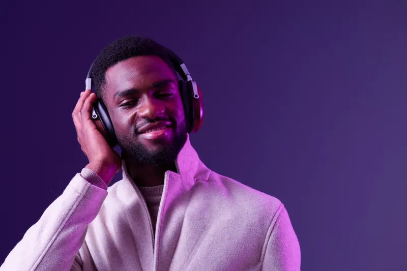 A young Black man enjoying music with headphones in a stylish outfit, featuring a modern purple background, conveying relaxation and joy through music