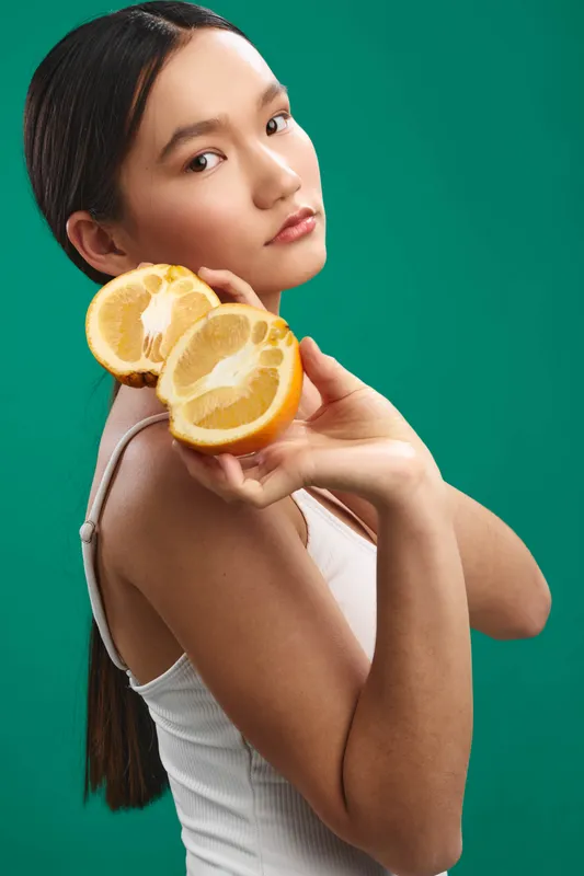 asian woman holds citrus slices close to her face in a vivid studio setting. she gazes softly, projecting a fresh and energetic mood and a modern lifestyle vibe.