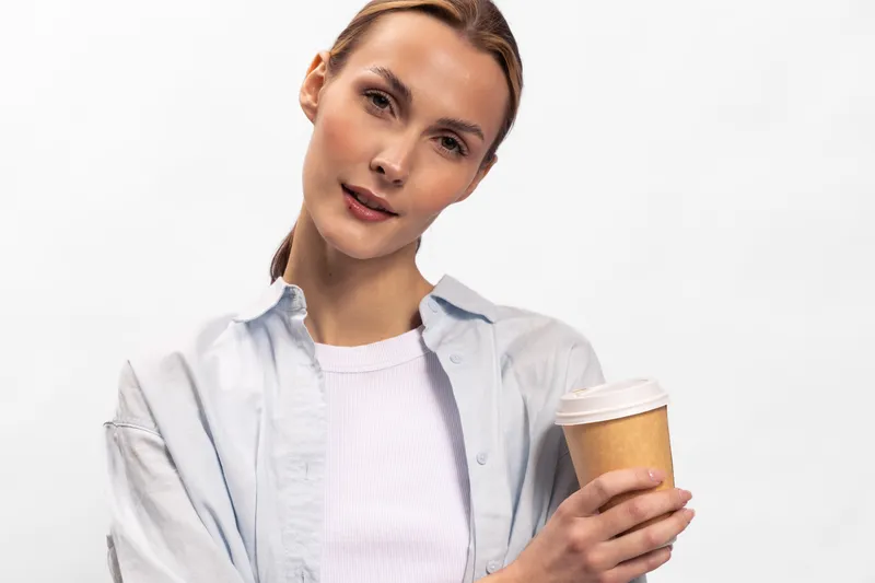 Young woman with light skin and blonde hair holds disposable coffee cup, wearing casual white shirt and light jacket. Studio portrait with soft lighting on white background.
