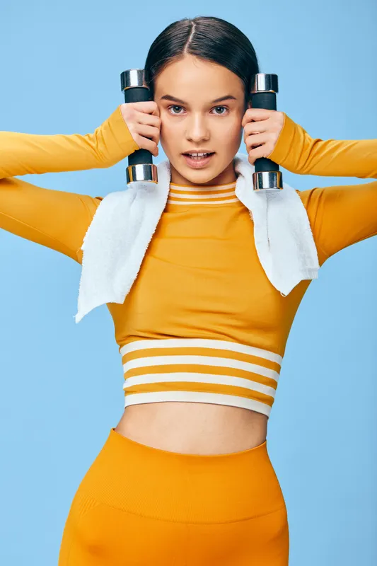 Fitness scene with a focused woman in bright orange activewear lifting dumbbells, a towel draped over shoulders, against a blue backdrop, capturing strength and athletic dedication.