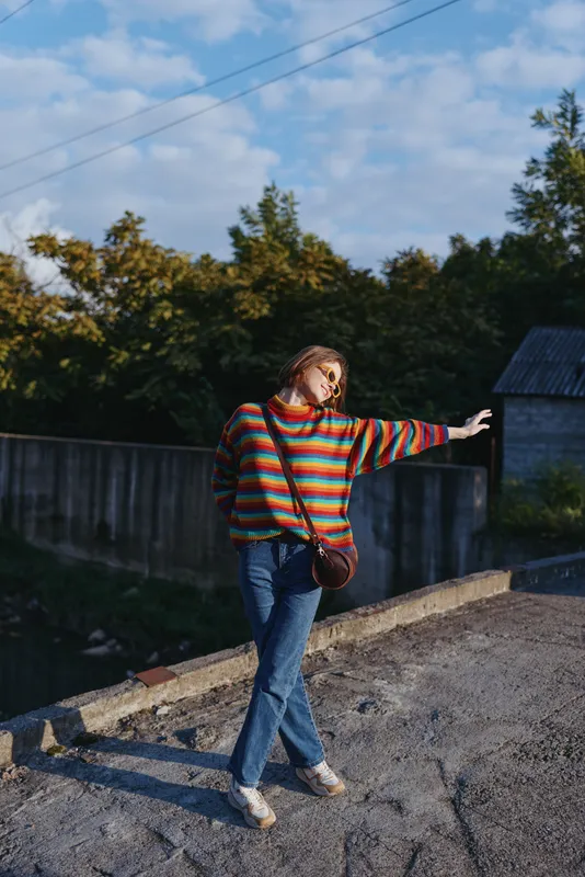 Woman in a colorful sweater and jeans posing outdoors on a rural rooftop with a crossbody bag, playful pose in warm sunlight for casual fashion, lifestyle portrait and seasonal street style.
