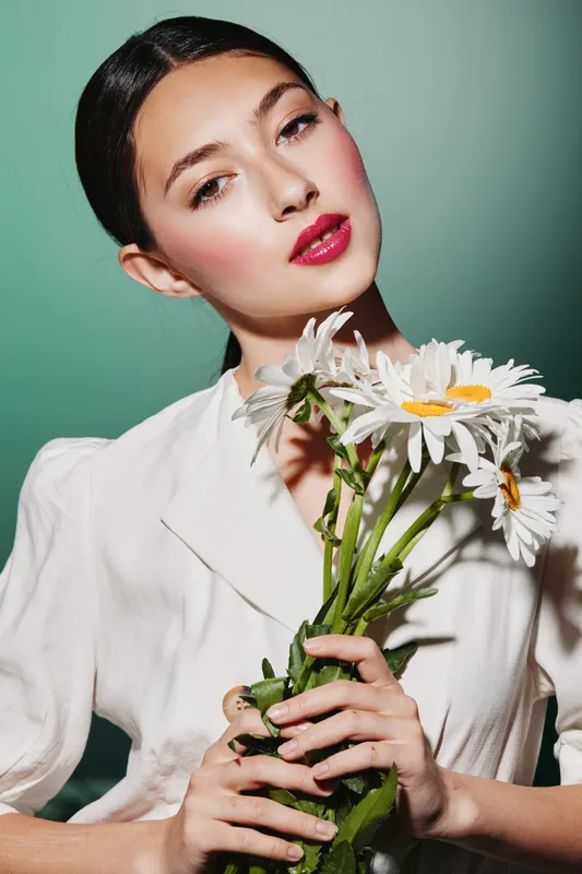 Elegant young woman with glossy dark hair and bright red lips holds white daisies. Fashion beauty close-up on green background emphasizes fresh glamor and softness.