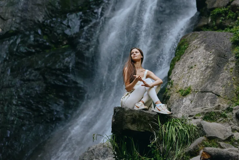 Young woman sitting on rock near waterfall in nature, wearing white activewear and sneakers, enjoying peaceful outdoor moment with calm expression and long hair.