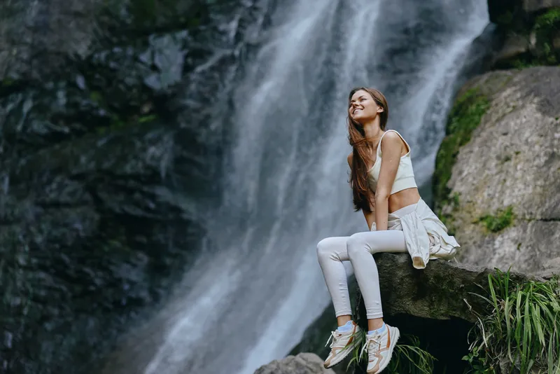 young woman sitting near waterfall in white activewear, enjoying nature and sunlight with relaxed and happy expression during outdoor adventure