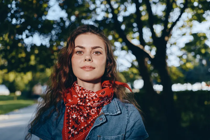 Lifestyle woman in modern boho-western denim street style with film color effect, outdoors in natural sunlight, wearing a red bandana and casual blue jacket.