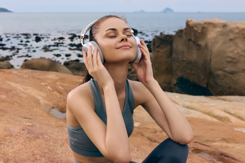 Young woman enjoying music by the sea in activewear, smiling blissfully with headphones on, surrounded by rocky beach landscape